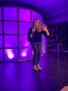 A photo of Ceilidh Henderson, a white woman with blond hair standing in front of a purple lit wall. She is wearing a black top and grey pants. She is facing the camera with a shocked look on her face. Her left hand is raised to shoulder level with her hand spread and holding the microphone in her right hand as she is addressing the audience.