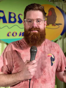 A photo of Chad Noonan, a white man with glasses and red hair, with a beard. He is wearing a pink t-shirt standing at a comedy club holding a microphone.
