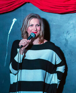 Photo of Heather Hurst. a white woman standing in front of a black wall at a comedy club wearing a white and black stripped top speaking into a microphone, performing a standup set.
