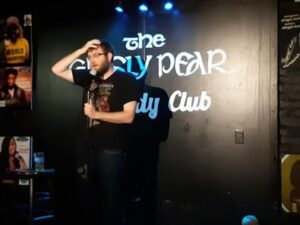 A photo of Matthew Champ, a white man with light brown hair, a beard and wearing glasses. He is performing at the Grisley Pear Comedy Club in NY. He is holding the mic in his left hand while talking into it and scratching his head with his right hand.
