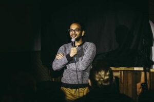 A photo of Tash Naved, a Middle Eastern man with black facial hair. He is wearing a grey sweater with black trim and olive green pants. His arms are crossed and he is talking into a microphone to the audience.