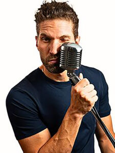 A photo of Rory Gardiner, a white man holding a radio holding a micrphone by the stand pretending to punch himself in the face with it.