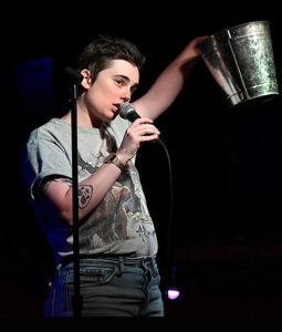 A photo of Sophie, a white woman wearing a grey short sleeved t-shirt. She is standing next to a mic stand., holding a mic in her right hand holding up a bucket with her left hand.