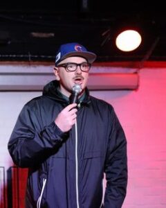 A photo of Tommy Fitz, a white man with a mustache and wearing glasses. He is wearing a Chicago Cubs baseball cap and a navy windbreaker. He is talking into a microphone to the audience.