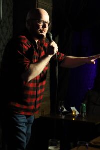 There is a photo of Zachariah, a white bald man with a red beard, wearing a red and black checkered shirt talking into a microphone in his right hand while holding his left hand out to the audience.