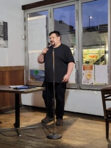 A photo of Hamilton Burger, a white male with brown hair and a goatee. He is wearing a black t-shirt and black pants as he performs holding the microphone in his right hand and with his left arm resting by his side.