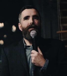 A photo of Hart Shouldice, a white man with black hair and a beard. Hart is holding a microphone in his left hand speaking into it.