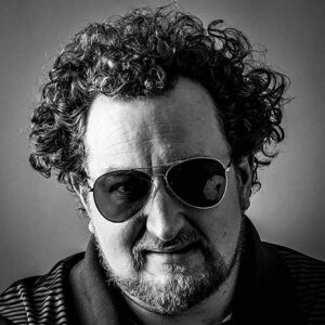 A black and white photo of Kris Kingsbury, a white man with curly hair, a beard and wearing sunglasses.