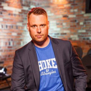 Photo of Lewis Hill, a white man in front of a blurred brick wall, wearing a t-shirt and grey blazer.