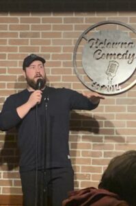 A photo of Nick Gent, a white man with brown hair and a beard. He is wearing a black cap and a black top. He is performing at Kelowna Comedy. Nick is speaking into the mic he is holding with his right hand while motioning with his left hand.